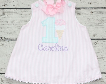 May include: Pink and white striped dress with a large pink bow at the top. The dress features a number 1 and an ice cream cone applique, with the name "Caroline" embroidered below. The dress has a scalloped hem.