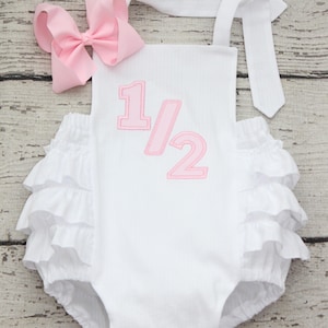May include: A white ribbed romper featuring a pink embroidered 1/2 applique on the front, multiple layers of ruffles on the sides, and a matching pink hair bow. The garment has white shoulder straps that tie at the top for an adjustable fit.