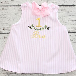 May include: Pink and white striped baby dress with a large pink bow at the neckline. The dress features yellow embroidered details, including the number "1", the name "Bea", and bee and leaf accents.