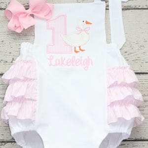 May include: White baby romper with pink accents, featuring a pink bow, a large pink number 1, and a duck applique. The name "Lakeleigh" is embroidered in pink. The romper has ruffled trim and striped fabric.