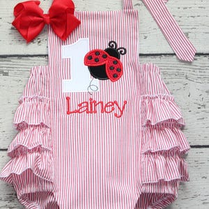 May include: A red and white striped romper featuring a large white number one, a red and black ladybug appliqué, and the embroidered name Lainey in red script. The garment includes three tiers of ruffles on each side and a matching red hair bow.