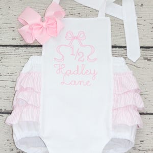 May include: A white romper featuring pink striped ruffles on the sides and a pink embroidered design on the front that includes a bow, the fraction 1/2, and the name Hadley Lane. A matching pink hair bow is attached to the top left strap.