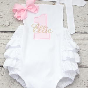 May include: White baby romper with a pink number one and the name "Ellie" embroidered in gold. The romper has ruffled trim and a large pink bow. The straps tie at the shoulders.
