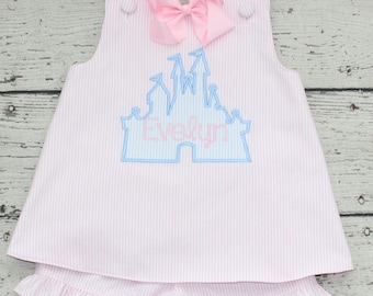 Può includere: Un completo composto da top senza maniche e pantaloncini con volant, caratterizzato da un motivo a righe verticali rosa chiaro e bianco. Il top presenta il contorno di un castello ricamato in azzurro con il nome Evelyn in rosa e un fiocco.