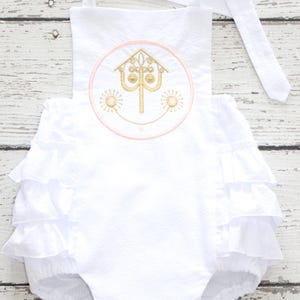 May include: White baby romper with a ruffled bottom and a round embroidered design. The design features a gold-colored building with a smiling face and two suns, outlined in pink. The romper has white straps.