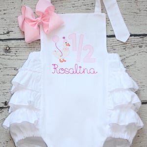 May include: White baby romper with pink embroidered details. Features a pink bow, ruffles, and the text "1/2 Rosalina" with a goose holding a balloon. The romper has tie straps.