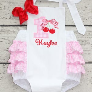 May include: White romper with pink gingham ruffles and a red bow. The romper features a pink gingham number 1 with a cherry design and the name "Kaylee" embroidered in red. The straps are white.