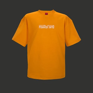 Op de afbeelding: Een oranje T-shirt met korte mouwen en witte Cyrillische tekst over de borst. Het shirt heeft een ronde hals en een klein rood label bij de halslijn. De achtergrond is donkergrijs.