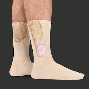 May include: A pair of beige socks with a humorous design. One sock features a brown and tan graphic, while the other has a pink oval shape. The socks are made of a soft, comfortable material.