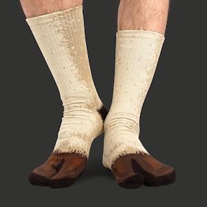 May include: A pair of novelty socks designed to resemble deer hooves. The socks are primarily beige with brown accents at the toes and heels, and a darker brown color for the hoof design. The socks are ribbed and extend up the calf.
