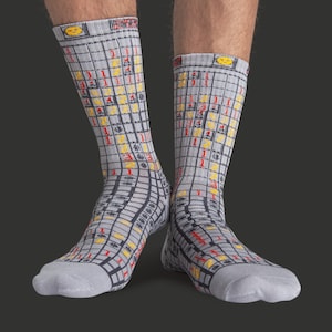 Op de afbeelding: Een paar grijze sokken met een rasterpatroon met rode, gele en zwarte details. De sokken hebben een smiley-gezichtontwerp aan de bovenkant. Een leuk en uniek accessoire.