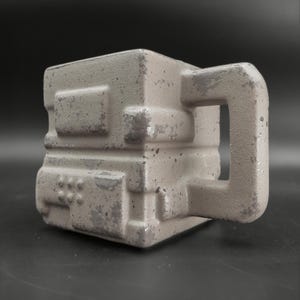 Peut inclure: Une tasse carrée gris clair avec un design géométrique et une anse carrée assortie. La tasse a une surface texturée avec des accents gris plus foncés et un aspect moucheté, suggérant un matériau en béton ou en pierre.