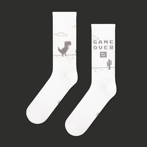 May include: White crew socks featuring a pixelated dinosaur and the words "GAME OVER." The socks have a retro video game theme, with a desert landscape and cactus design. The socks are a fun and playful accessory.