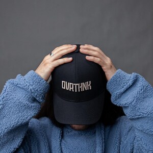 Könnte beinhalten: Eine marineblaue Baseballkappe mit dem Wort "OURTHNK" in Silber gestickt. Die Kappe wird von einer Person getragen, die eine blaue Fleecejacke trägt. Der Hintergrund ist einfarbig grau.