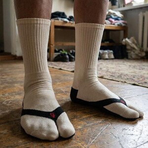 May include: Cream-colored socks with a black thong-style strap and a red emblem. The socks have a ribbed texture and are designed to separate the big toe. The socks are worn on a wooden floor.