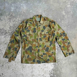 May include: A camouflage jacket in shades of green, brown, and grey. The jacket features a button-up front, two chest pockets, and long sleeves. The fabric has a mottled pattern, and the jacket appears to be vintage or military-inspired.