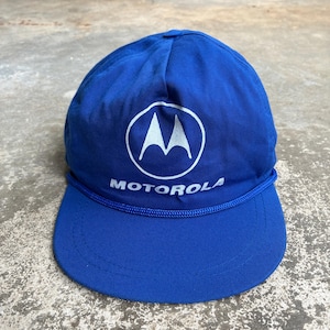 May include: A bright blue baseball cap featuring the white Motorola logo and brand name printed on the front. The cap includes a matching blue braided cord trim across the base of the crown, just above the stiff, curved brim of the hat.