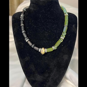 Puede incluir: Un collar artesanal exhibido en un busto de terciopelo negro. El collar presenta una mezcla de cuentas negras, plateadas y verdes. Una gran cuenta dorada con un diseño floral está centrada en el collar.