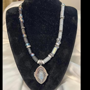 May include: A handcrafted necklace with a silver pendant and a beaded design. The pendant is oval-shaped with a light blue stone. The necklace features a mix of black, silver, and iridescent beads. The necklace is displayed on a black mannequin.