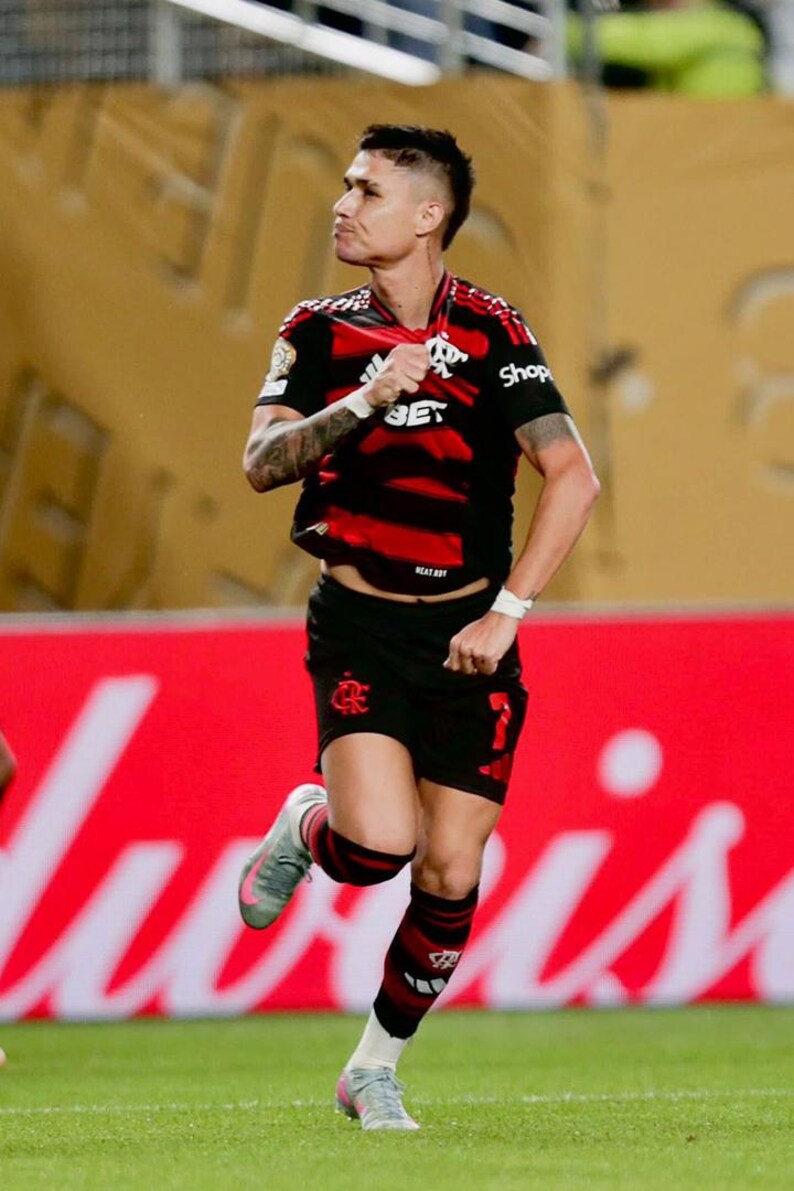 Puede incluir: Un futbolista con una camiseta a rayas negras y rojas y pantalones cortos negros corre por un campo verde. La camiseta tiene un logo y la palabra "BET". El jugador lleva zapatos grises y rosas y calcetines a rayas negras y rojas.