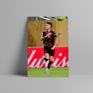 Puede incluir: Impresi&oacute;n enmarcada de un jugador de f&uacute;tbol con camiseta a rayas negras y rojas y pantalones cortos negros, corriendo sobre un campo verde. El fondo incluye un letrero rojo con texto blanco y una pared amarilla.
