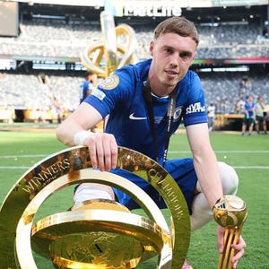 May include: A soccer player in a blue jersey kneels on a green field, holding a large gold trophy. The trophy has a globe design and the words "WINNERS" and "CHAMPIONS" are visible. A smaller gold trophy is held in the other hand.