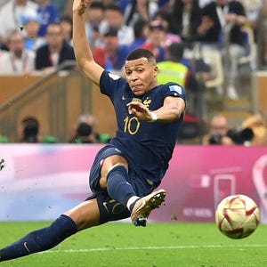 May include: A soccer player in a navy blue uniform with the number 10 kicks a soccer ball on a green field. The player is mid-kick, with his leg extended and arm raised. The background shows a crowd of spectators.