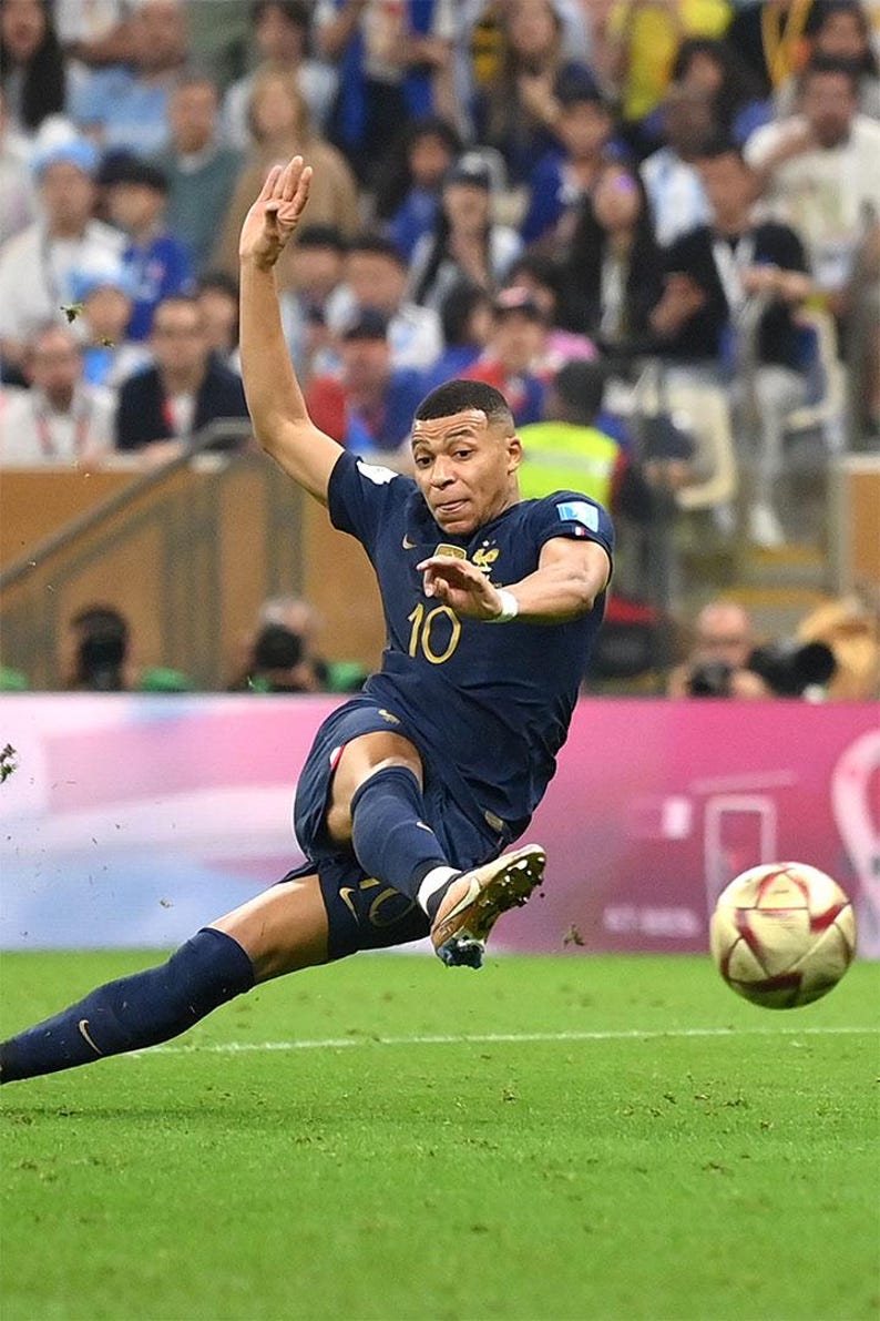 May include: A soccer player in a navy blue uniform with the number 10 kicks a soccer ball on a green field. The player is mid-kick, with his leg extended and arm raised. The background shows a crowd of spectators.