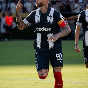 May include: A soccer player in a navy and white jersey with the word "codere" on the front, number 93 on the shorts, and red socks. The player is on a green field, raising a finger.