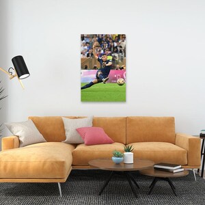 May include: A framed print of a soccer player in a blue uniform kicking a soccer ball on a green field. The player is mid-kick, with the ball in front of them. The background shows a crowd of spectators.