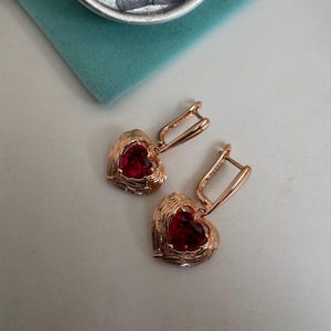 Puede incluir: Un par de pendientes en forma de corazón de oro rosa con un centro de piedra roja. Los pendientes tienen un diseño texturizado, similar a un tejido, alrededor del corazón. Se exhiben sobre una superficie blanca, con una bolsa de terciopelo verde azulado y una moneda de plata en el fondo.