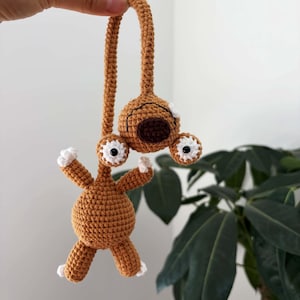 Pode incluir: Uma criatura de crochê feita à mão em tons de castanho, com detalhes brancos nos olhos e nos pés. O design caprichoso apresenta um corpo redondo, braços estendidos e uma alça para pendurar. Está pendurado em frente a uma planta verde e frondosa.