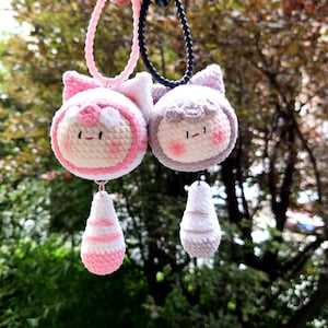 May include: Two handmade crochet pendants. One is pink and white, the other is gray and white. Each pendant features a round face with rosy cheeks, small eyes, and a bow. The pendants are attached to a pink and black cord.