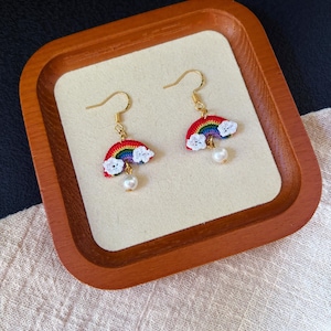 May include: A pair of gold-toned hook earrings featuring a small, multi-colored crocheted rainbow charm. Each rainbow is accented with two white crocheted clouds at the ends and a single dangling white faux pearl bead suspended from the center.
