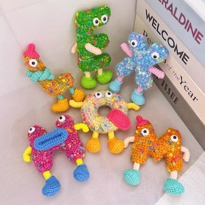 Op de afbeelding: Een verzameling van vijf gehaakte pluche figuren met textuur in de vorm van letters en een donut. Elk figuur heeft wiebeloogjes, kleine ledematen en is gemaakt van levendig garen met veelkleurige spikkels. De set bevat lettervormen en een donut.