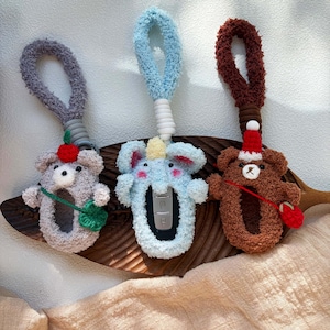 May include: Three animal-shaped keychains. The keyrings are grey, blue, and brown, featuring a bear, elephant, and bear design. Each has a loop for carrying. The bear keychain has a red hat and mitten. The keyrings are made of plush material.
