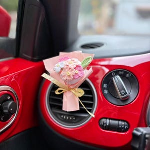 Könnte beinhalten: Ein kleiner Strauß gehäkelter Blumen in Rosa, Weiß und Grün, verpackt in rosa Papier und mit einem goldenen Band gebunden. Der Strauß befindet sich in einem roten Autoinnenraum in der Nähe der Lüftungsöffnung.