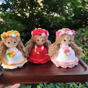 May include: Three handmade crochet dolls with long brown hair and floral headbands. The dolls wear white, red, and pink dresses with floral accents. The dolls are displayed on a wooden tray.