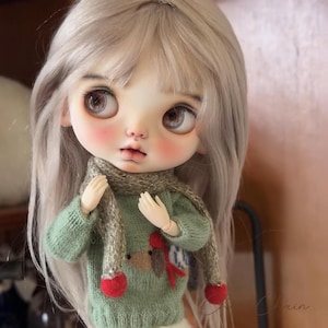 May include: A doll with large eyes and long, light gray hair. The doll wears a green sweater with a reindeer design and a gray scarf with red heart-shaped accents. The doll's cheeks are rosy.