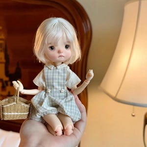 Könnte beinhalten: Eine kleine Gelenkpuppe mit kurzem weißem Haar und großen dunklen Augen. Die Puppe trägt ein weißes kurzärmeliges Hemd unter einem hellblau-weiß karierten Latzkleid mit kleinen sternförmigen Knöpfen. Sie hält einen kleinen geflochtenen Korb.