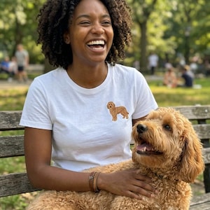 May include: A white short-sleeved t-shirt featuring a small, embroidered golden-brown doodle dog graphic on the left chest area. The shirt has a classic crew neckline and a simple, clean design, showcasing the detailed stitching of the dog illustration.