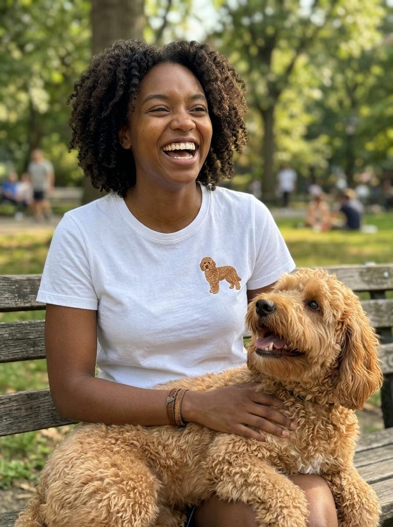 May include: A white short-sleeved t-shirt featuring a small, embroidered golden-brown doodle dog graphic on the left chest area. The shirt has a classic crew neckline and a simple, clean design, showcasing the detailed stitching of the dog illustration.