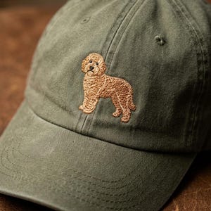May include: An olive green baseball cap featuring an embroidered design of a light brown, curly-haired dog standing in profile. The cap has a structured crown, a curved brim, and tonal stitching details throughout the fabric of the hat.