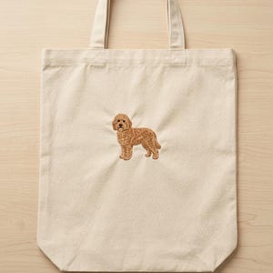 May include: A natural-coloured canvas tote bag featuring a centred, detailed embroidery of a golden-brown curly-haired dog standing in profile. The bag has two sturdy fabric handles at the top and a rectangular shape with a flat bottom base.