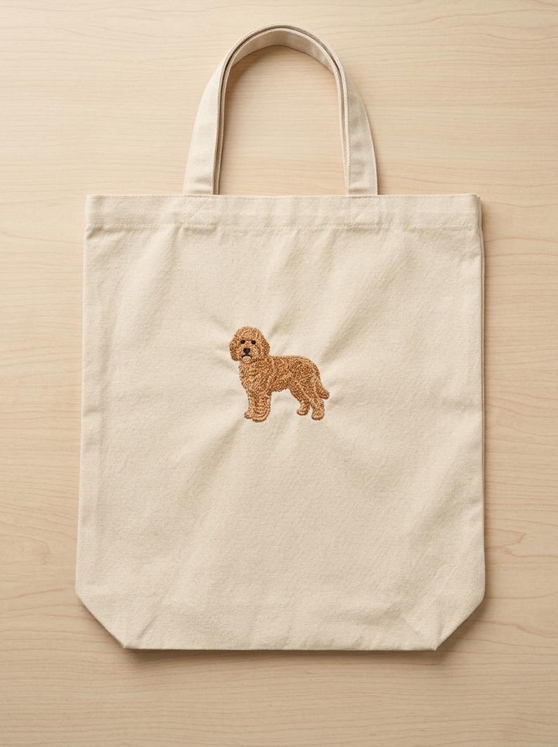 May include: A natural-coloured canvas tote bag featuring a centred, detailed embroidery of a golden-brown curly-haired dog standing in profile. The bag has two sturdy fabric handles at the top and a rectangular shape with a flat bottom base.