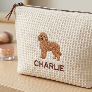 May include: A cream-coloured waffle-textured zipped pouch featuring an embroidered brown dog and the name CHARLIE stitched in brown thread below the dog. The pouch has a metal zip with a brown leather pull tab attached to the slider.