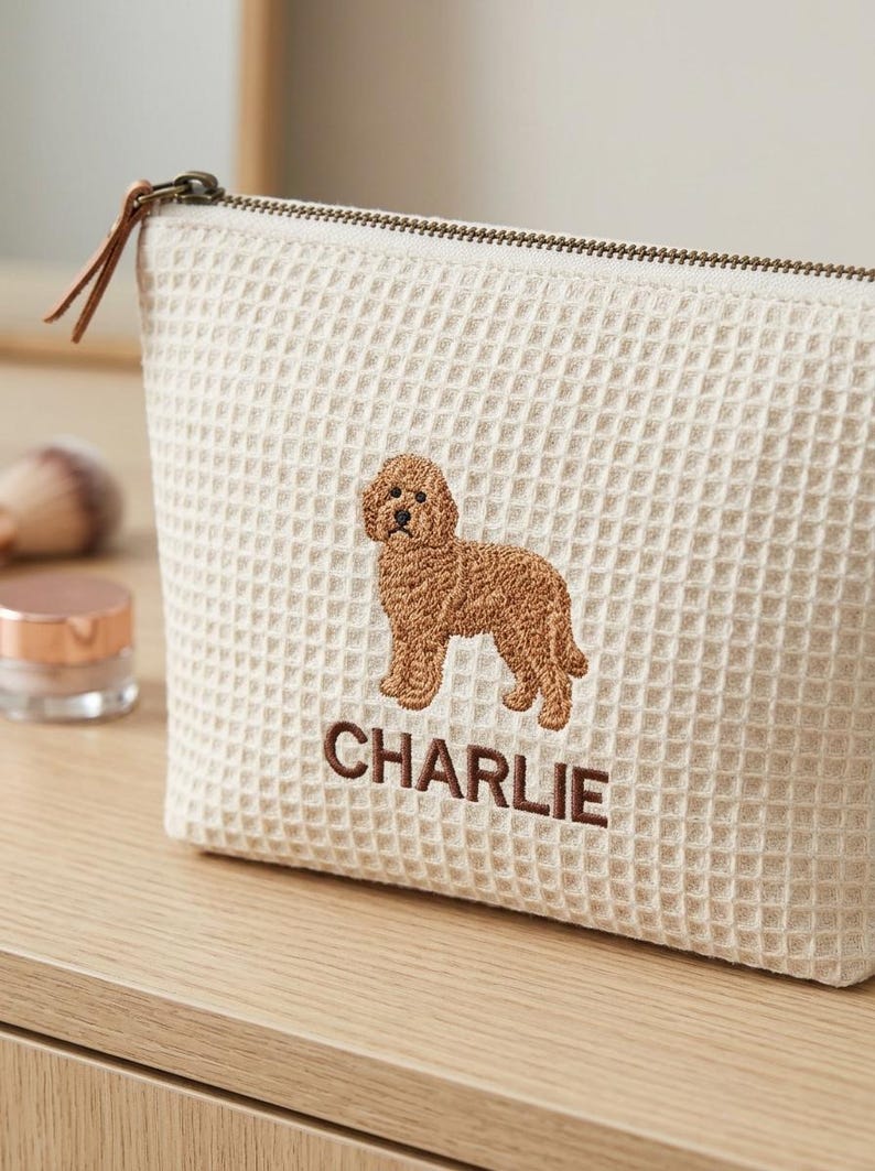 May include: A cream-coloured waffle-textured zipped pouch featuring an embroidered brown dog and the name CHARLIE stitched in brown thread below the dog. The pouch has a metal zip with a brown leather pull tab attached to the slider.