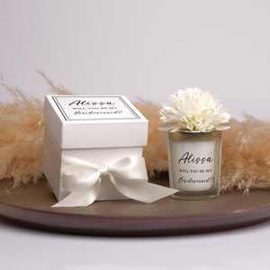May include: A white gift box tied with a satin bow, and a candle in a glass holder, both with the text "Alissa, Will You Be My Bridesmaid?" A white flower sits atop the candle. The items are on a brown tray.