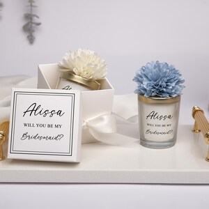 May include: A white gift box and a candle with the text "Alissa, Will You Be My Bridesmaid?" are displayed on a white tray. The box is tied with a white ribbon and topped with a white flower. The candle has a blue flower on top.
