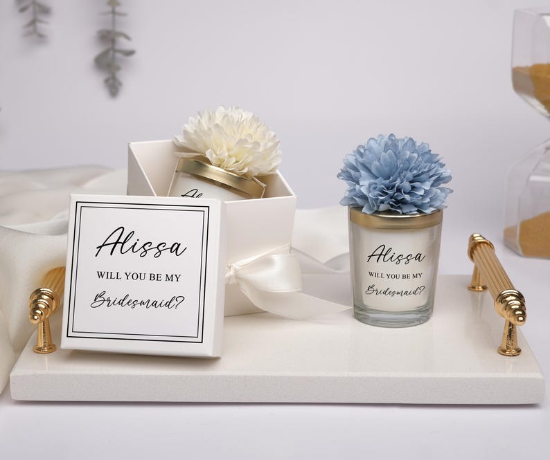May include: A white gift box and a candle with the text "Alissa, Will You Be My Bridesmaid?" are displayed on a white tray. The box is tied with a white ribbon and topped with a white flower. The candle has a blue flower on top.